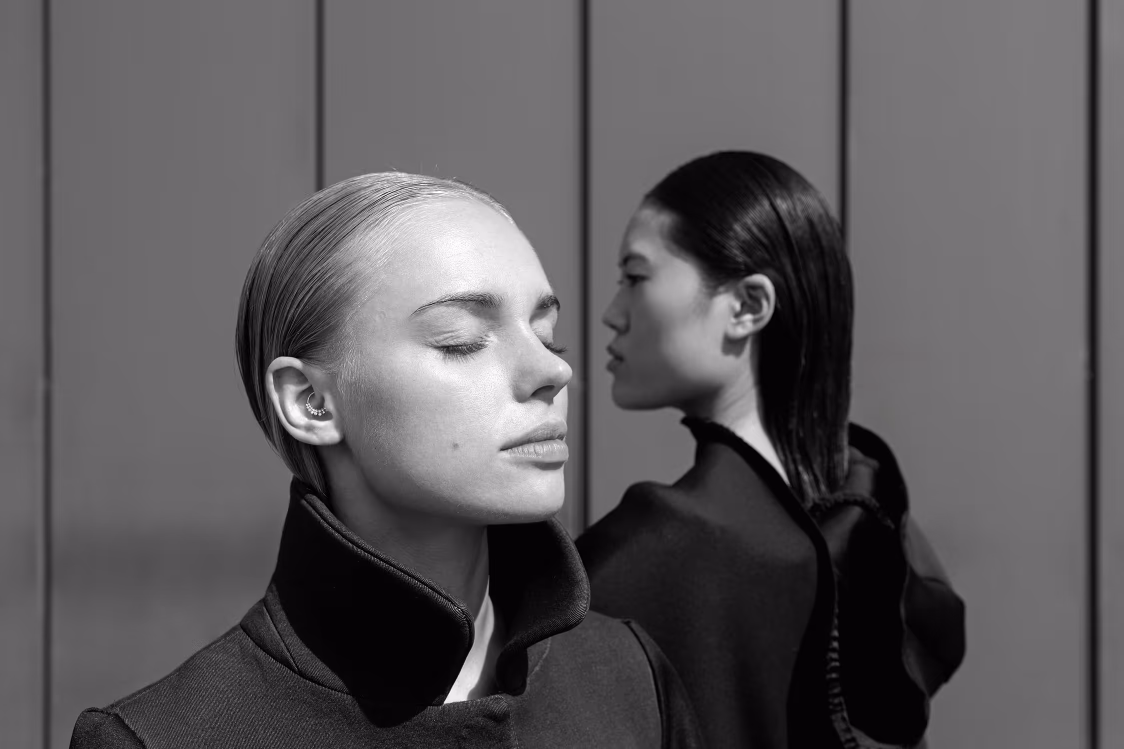 Two women standing by the wall, one of them has her eyes closed